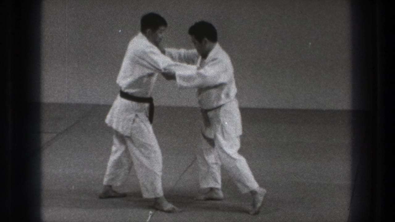 Two adult practitioners in white gi move laterally across an indoor mat in Tokyo, maintaining sleeve and lapel grips during judo practice.