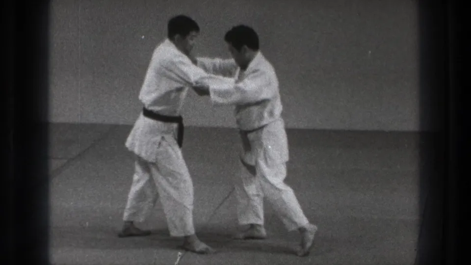 Two adult practitioners in white gi move laterally across an indoor mat in Tokyo, maintaining sleeve and lapel grips during judo practice.