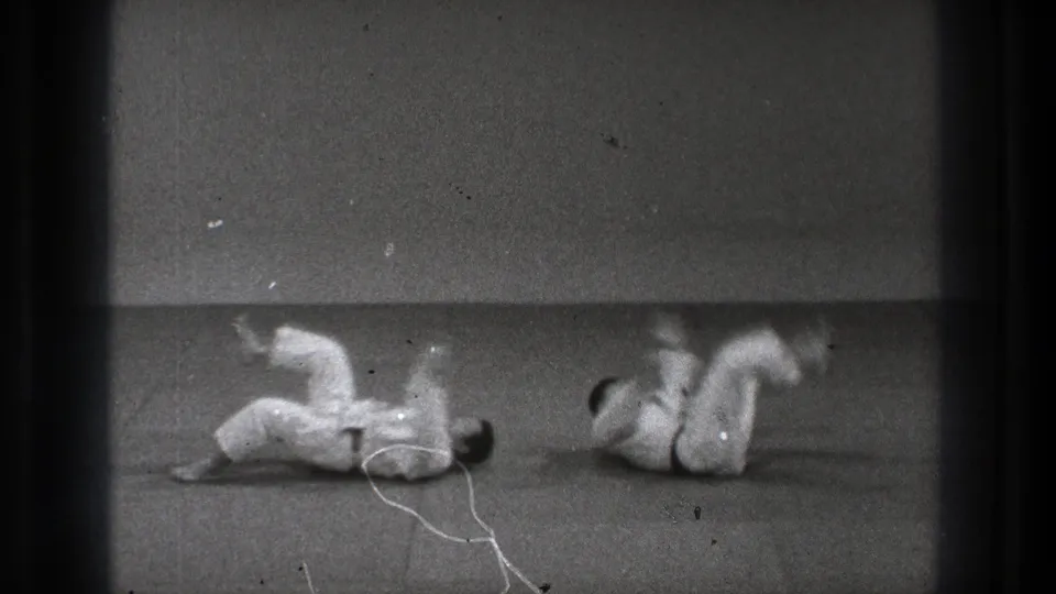 The throw sequence ends with both adult martial artists on the indoor mat, one rolling through the fall and the other landing nearby.