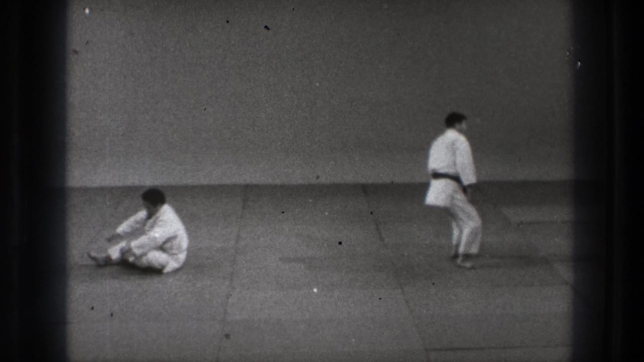 In 1971 Tokyo footage, one man in a white gi sits on the mat after a controlled fall as another stands across the sparse dojo.