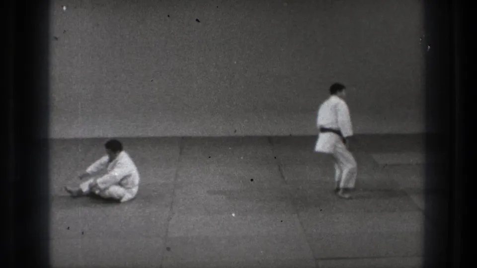 In 1971 Tokyo footage, one man in a white gi sits on the mat after a controlled fall as another stands across the sparse dojo.