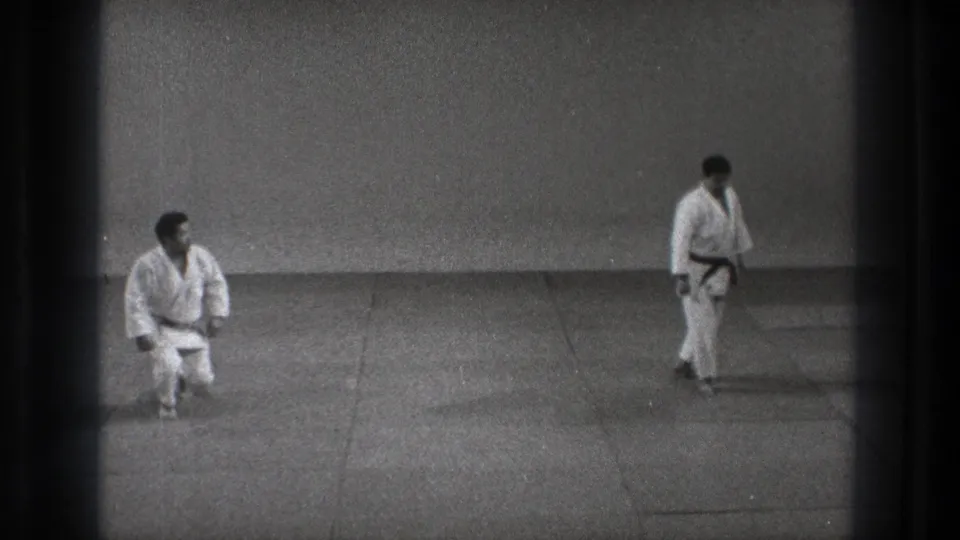 In the Tokyo dojo setting, one practitioner kneels after a movement while his partner in a white gi stands at a distance.