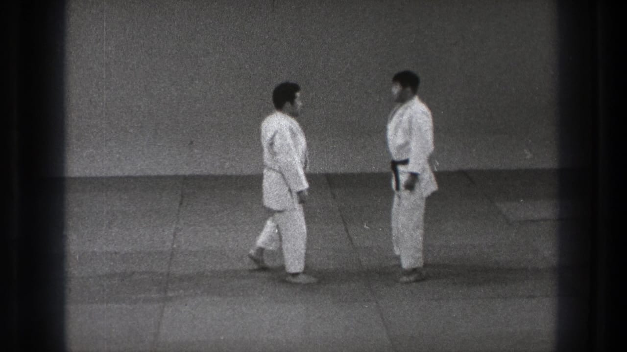 Two adult men in white judo uniforms face each other on a plain mat in supplied 1971 Tokyo footage.
