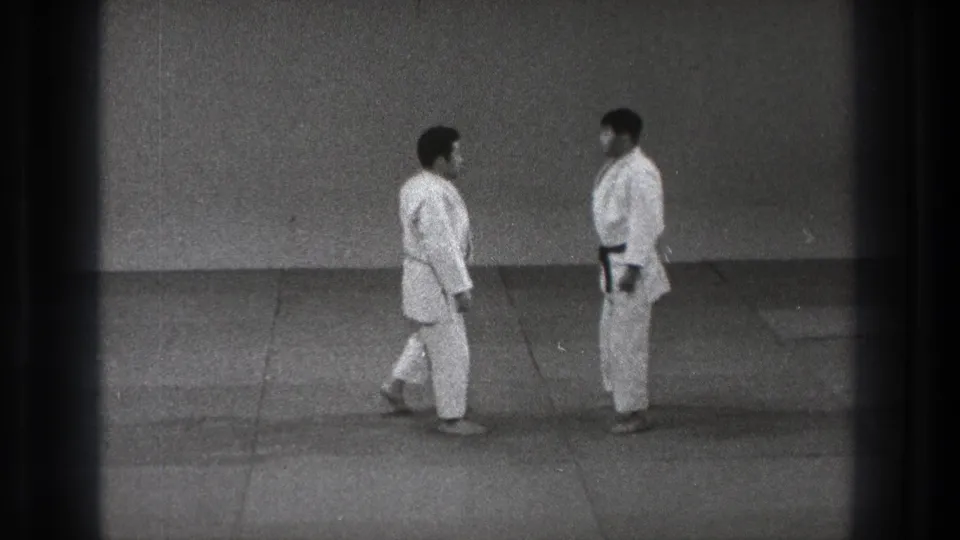Two adult men in white judo uniforms face each other on a plain mat in supplied 1971 Tokyo footage.