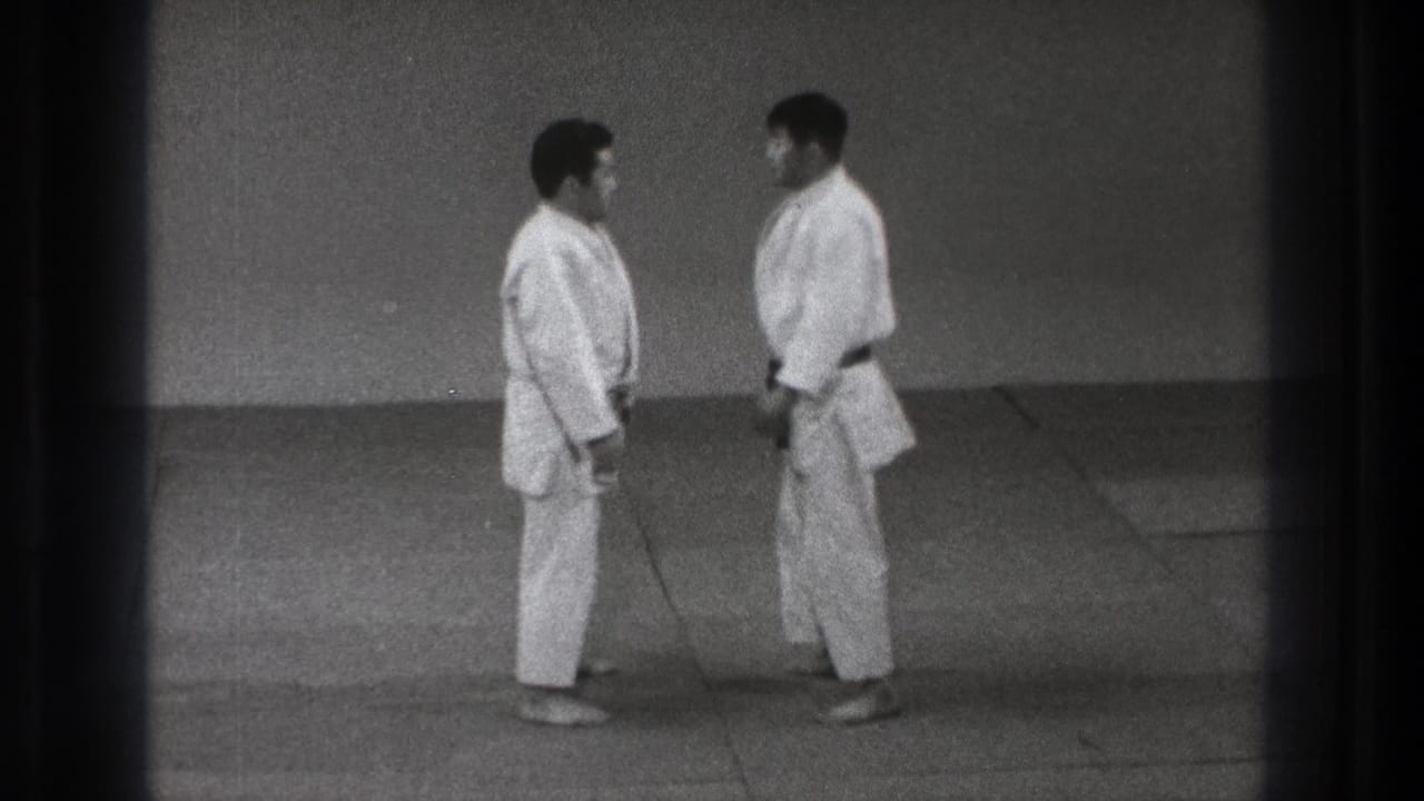 Two adult men in gi stand face to face on a mat, one wearing a dark belt, in archival Tokyo judo footage.