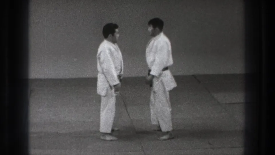Two adult men in gi stand face to face on a mat, one wearing a dark belt, in archival Tokyo judo footage.