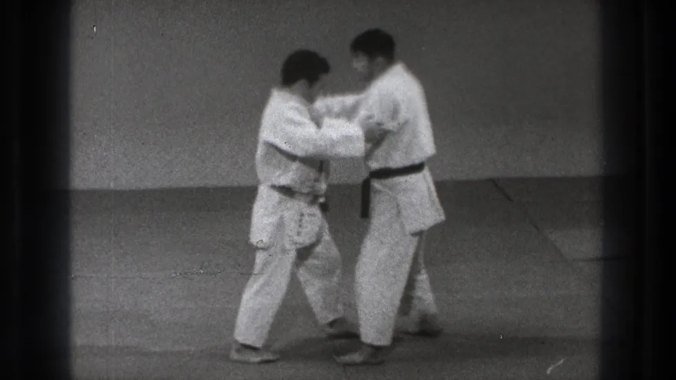Two judo practitioners begin a controlled grip exchange on the mat in the supplied 1971 Tokyo home-movie clip.