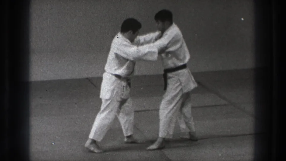 Two adult men in white gi and dark belts practice a standing grip on a mat in 1971 Tokyo footage.