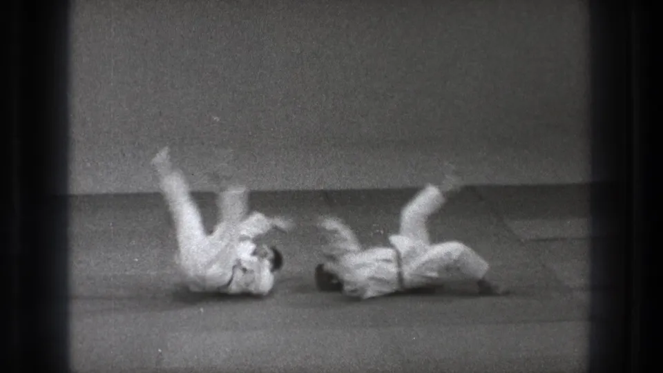 Two adult practitioners in white gi land on the mat during a judo-style throw in black-and-white 1971 Tokyo footage.