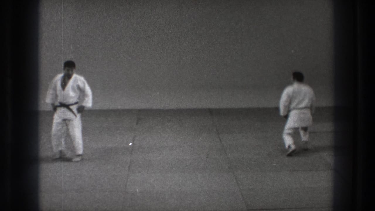 Two martial artists in white gi stand apart on a mat in a plain dojo.