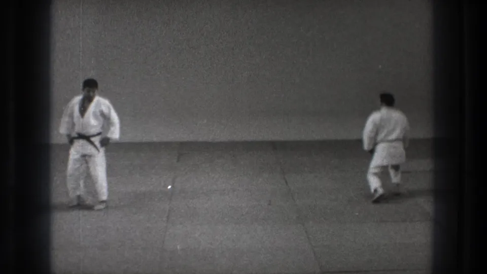 In 1971 Tokyo footage, two adult martial artists in white gi face each other across a mat in a simple indoor dojo.