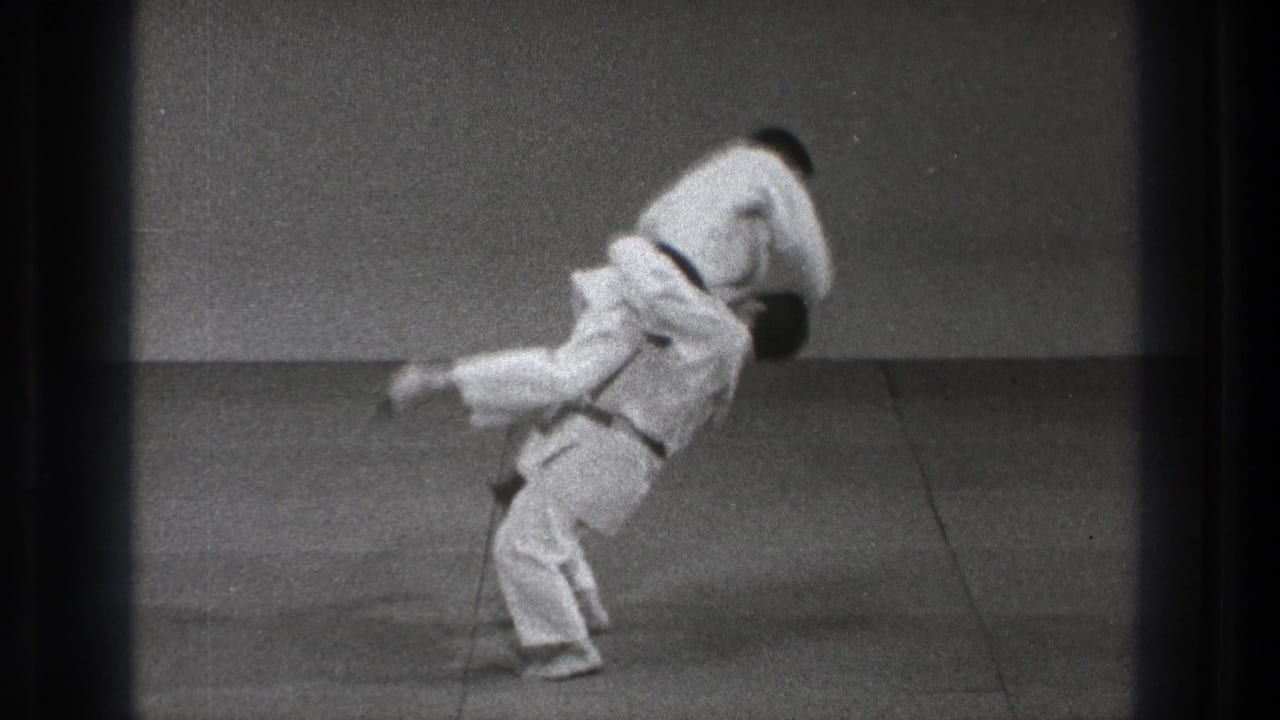 A judo-style throw is underway as one practitioner in a white gi lifts the other against the plain dojo wall and mat.