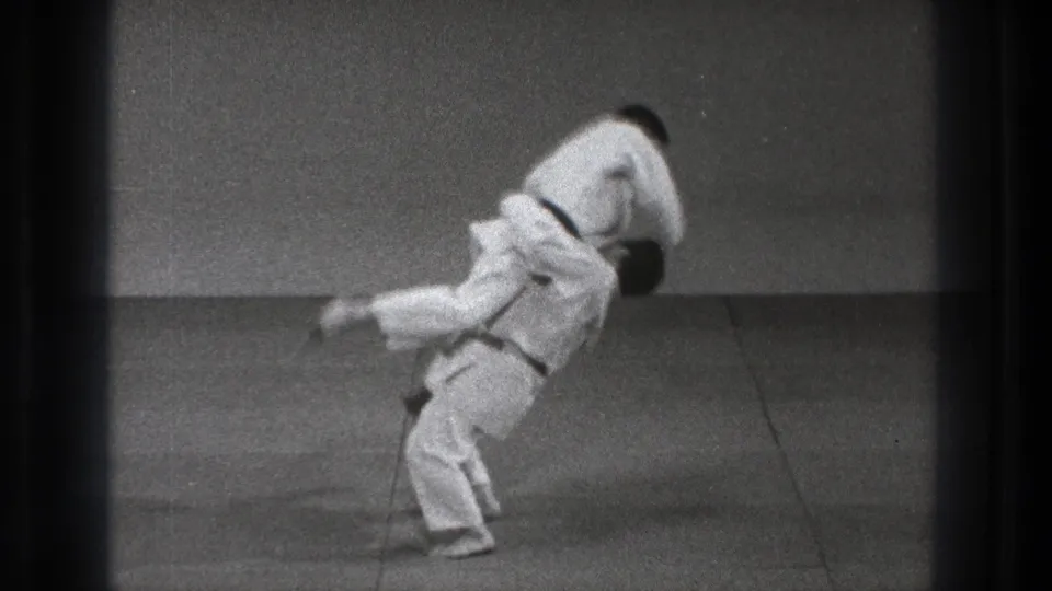 A judo-style throw is underway as one practitioner in a white gi lifts the other against the plain dojo wall and mat.