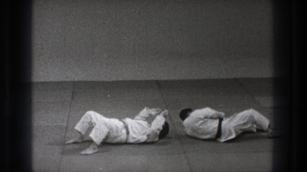 After the throw, both practitioners in white gi are down on the mat inside the simple Tokyo dojo setting.