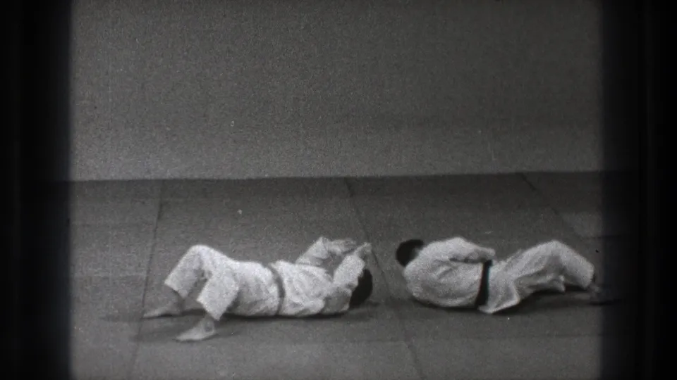 After the throw, both practitioners in white gi are down on the mat inside the simple Tokyo dojo setting.