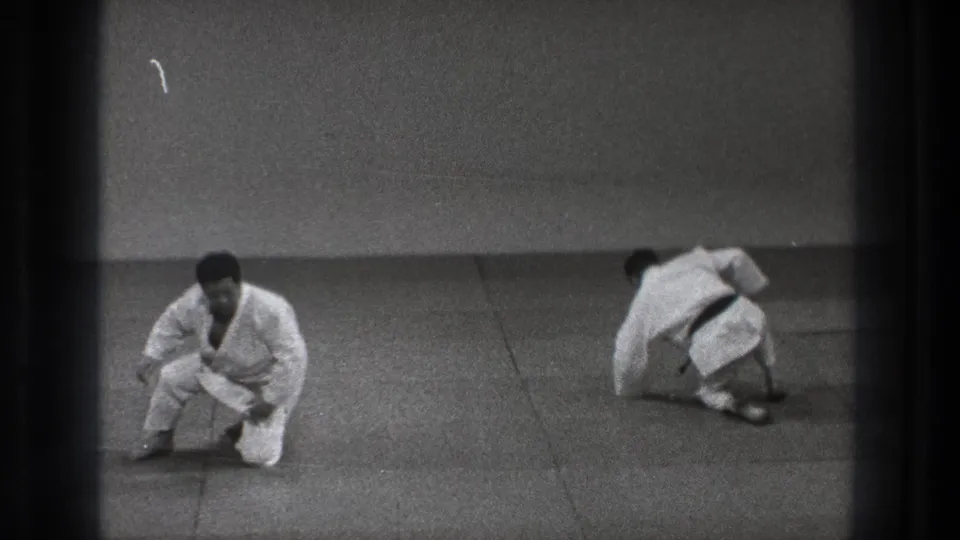1971 Tokyo 8mm frame showing one martial artist kneeling while the other turns on the mat during practice.