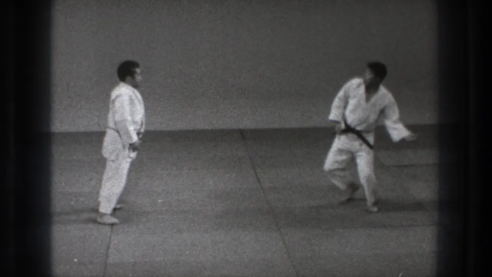 1971 Tokyo 8mm frame showing two martial artists in a dojo-like mat room, one standing while the other steps away.