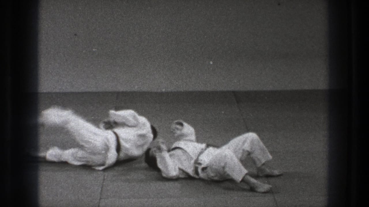 The judo demonstration reaches the landing phase as both practitioners fall and roll across the mat in the Tokyo 1971 home-movie footage.