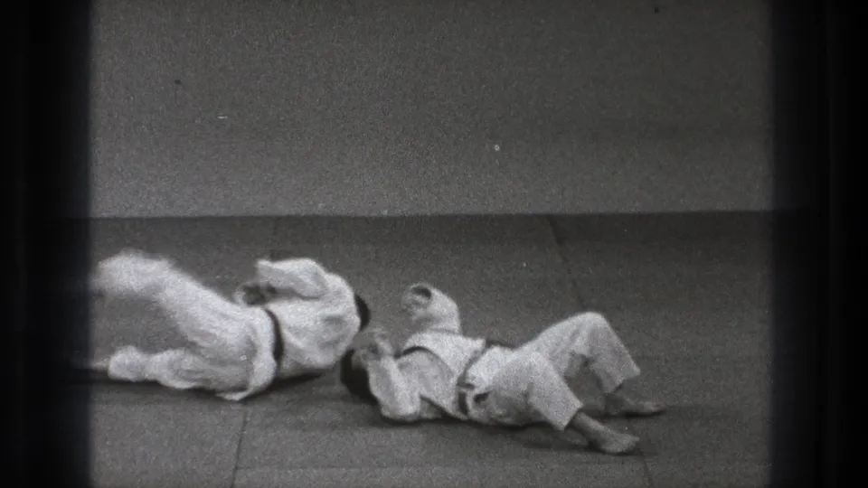 The judo demonstration reaches the landing phase as both practitioners fall and roll across the mat in the Tokyo 1971 home-movie footage.