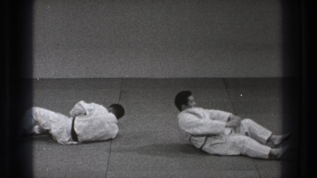 After the throw, the two black-belt practitioners remain low on the mat, one partly cropped at the edge and the other sitting back from the fall.