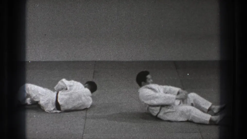 After the throw, the two black-belt practitioners remain low on the mat, one partly cropped at the edge and the other sitting back from the fall.