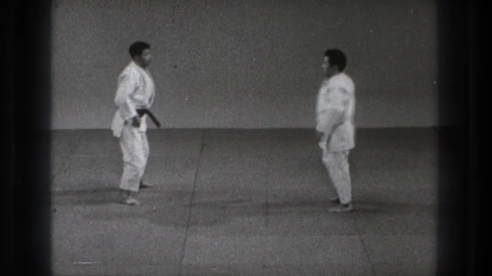 Two karate practitioners in white gi face one another in a simple indoor dojo setting in 1971 Tokyo footage.