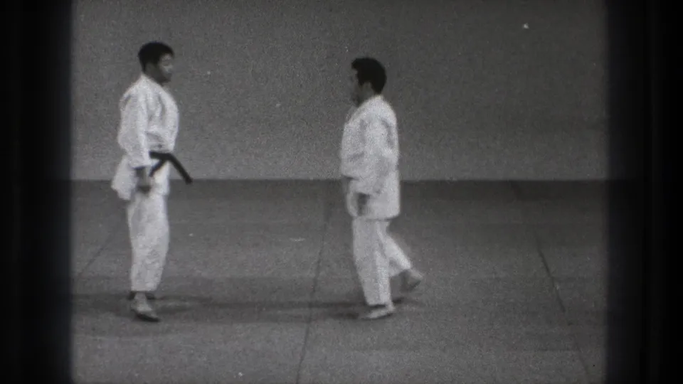 Two men in white karate uniforms walk toward one another on a tatami-style mat in archival Tokyo footage.