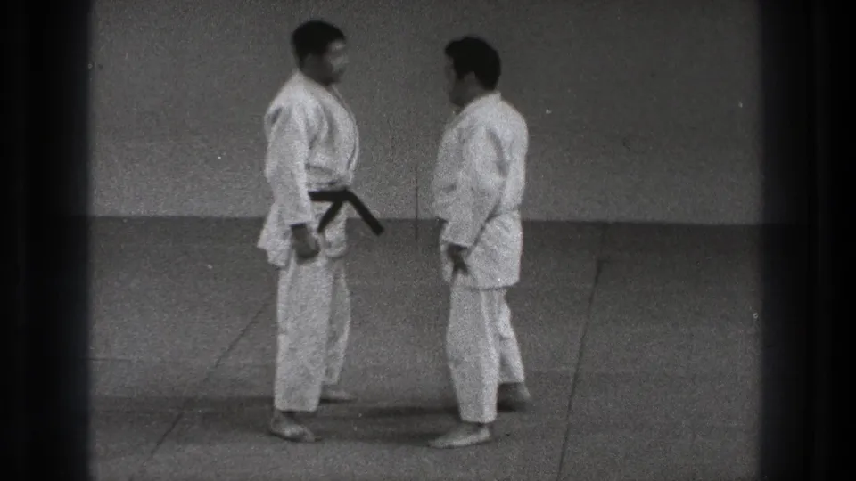 Two men in karate gi stand face to face at close range in a plain indoor practice space, Tokyo, 1971.