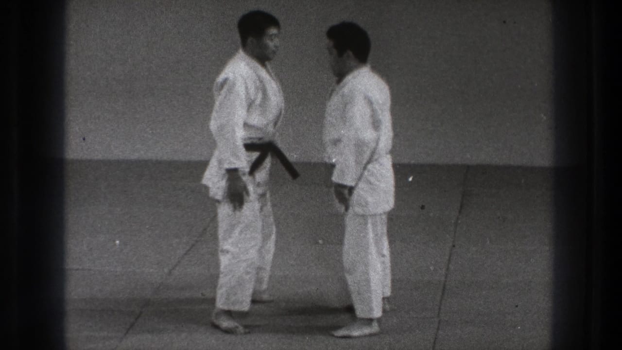 Two men in karate gi face each other on a mat in a plain training room.