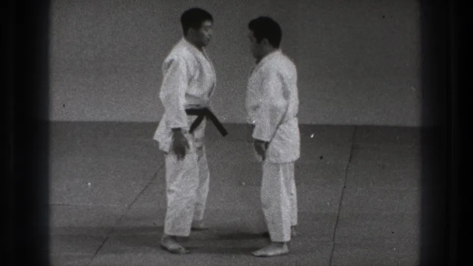 In Tokyo 1971 footage, two adult men in white karate gi stand barefoot on mats before beginning a technique demonstration.