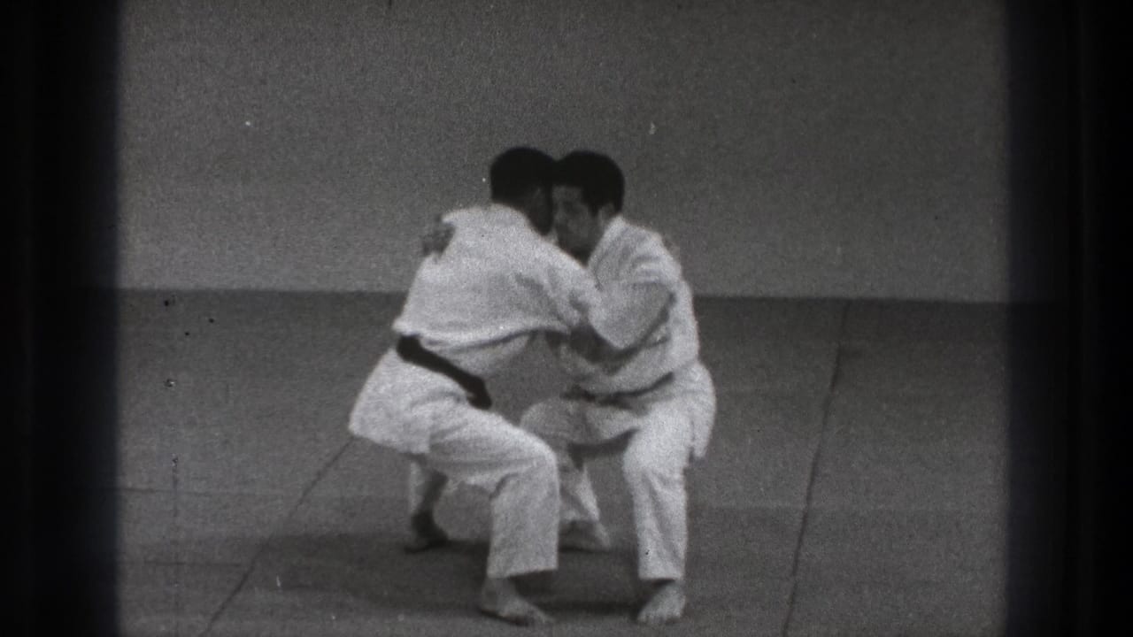 The men move into a close grappling exchange on the dojo mat, with the darker-belted practitioner controlling the position.