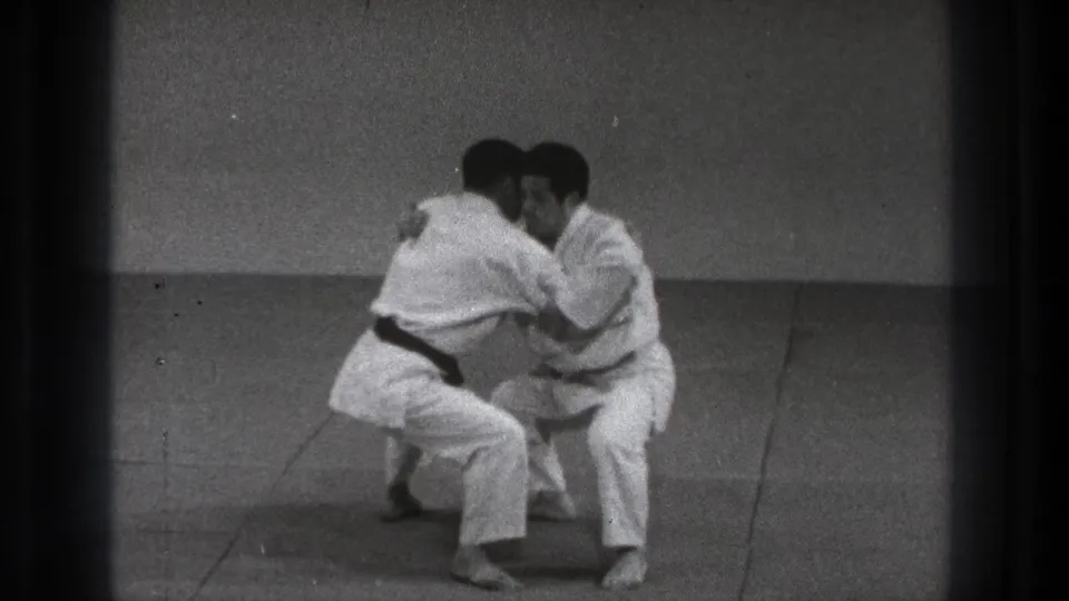The men move into a close grappling exchange on the dojo mat, with the darker-belted practitioner controlling the position.