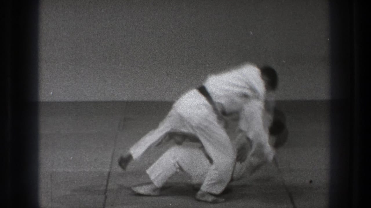 In a 1971 Tokyo martial arts demonstration, one man in a white gi throws another onto the mat in a plain training room.