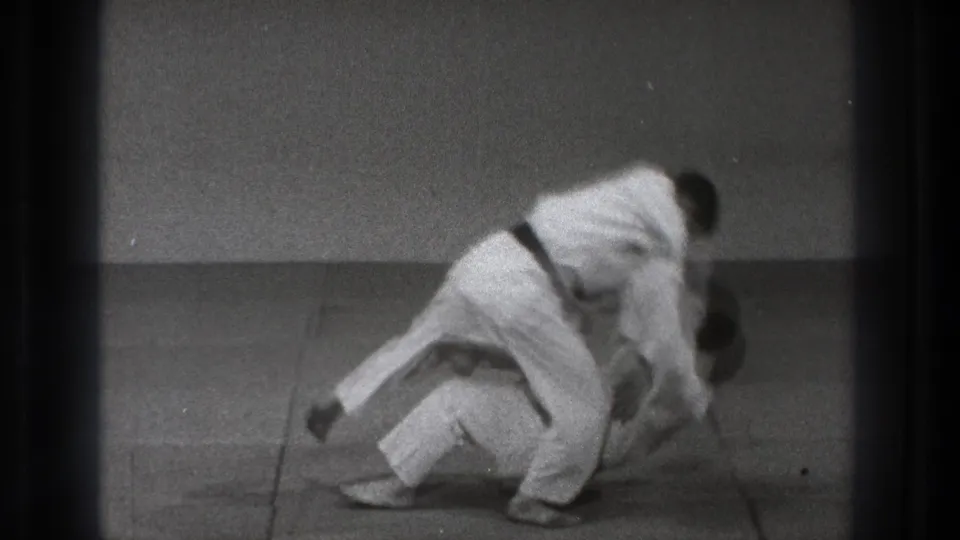 In a 1971 Tokyo martial arts demonstration, one man in a white gi throws another onto the mat in a plain training room.