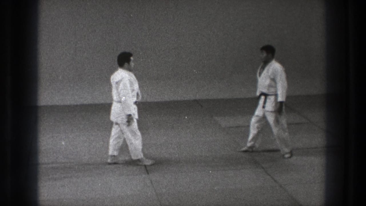 Two men in white gi approach each other across tatami mats in a plain dojo space.