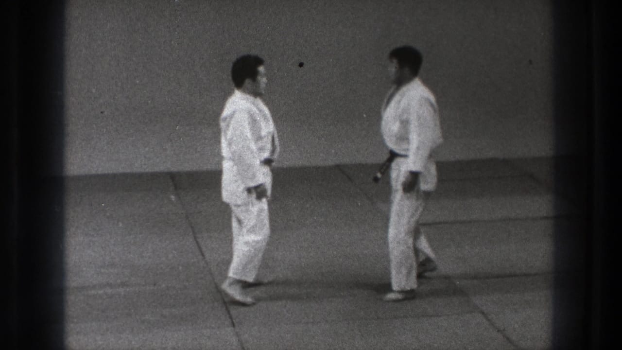 Two adult judo practitioners in white gi face each other at close range in the Tokyo dojo footage.