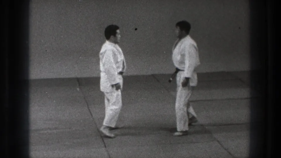 Two adult judo practitioners in white gi face each other at close range in the Tokyo dojo footage.