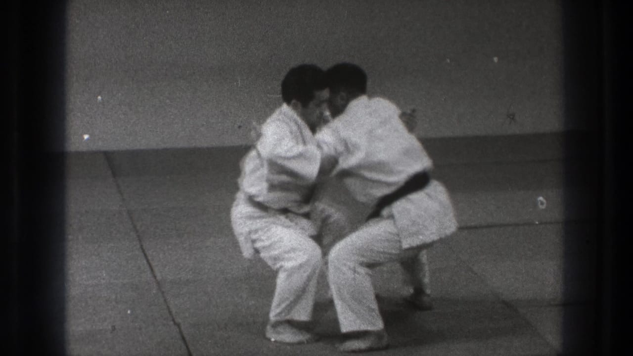 In 1971 Tokyo footage, two adult judo practitioners in white gi and black belts crouch in a close grip on tatami mats.