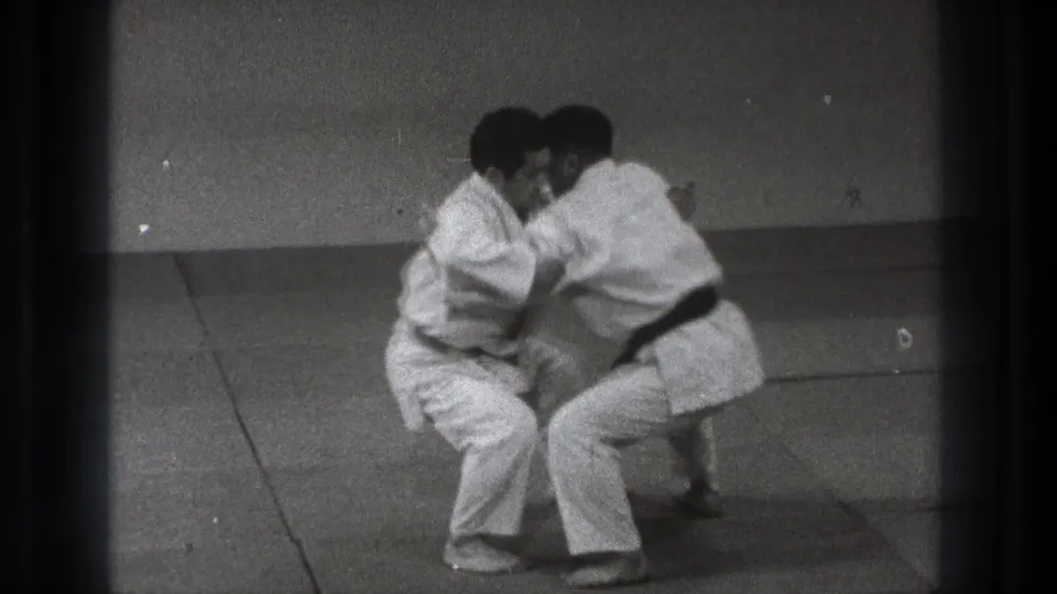 In 1971 Tokyo footage, two adult judo practitioners in white gi and black belts crouch in a close grip on tatami mats.