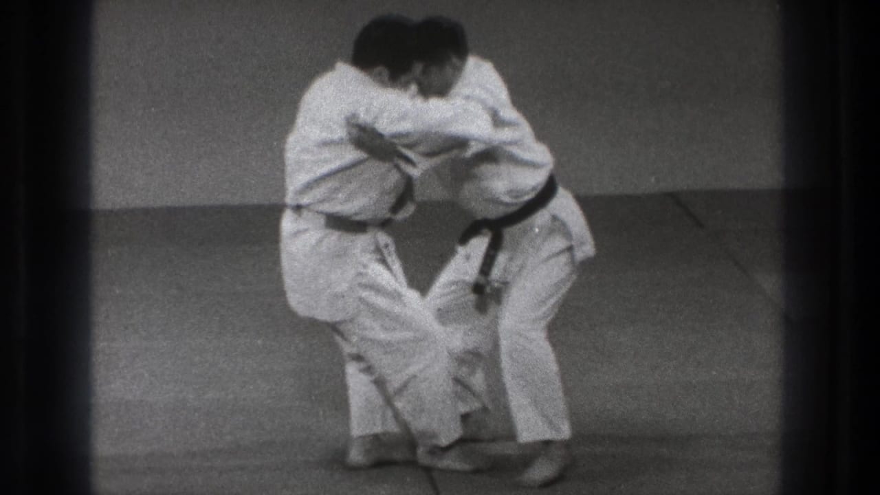Two adult judo practitioners in white gi and black belts stand chest-to-chest in a grappling hold in the Tokyo training space.