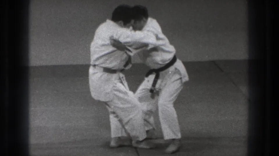 Two adult judo practitioners in white gi and black belts stand chest-to-chest in a grappling hold in the Tokyo training space.