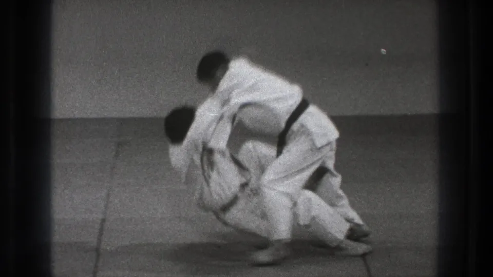 A judo throw is underway as one practitioner turns through the grip and the other loses balance over the tatami in the 1971 Tokyo clip.