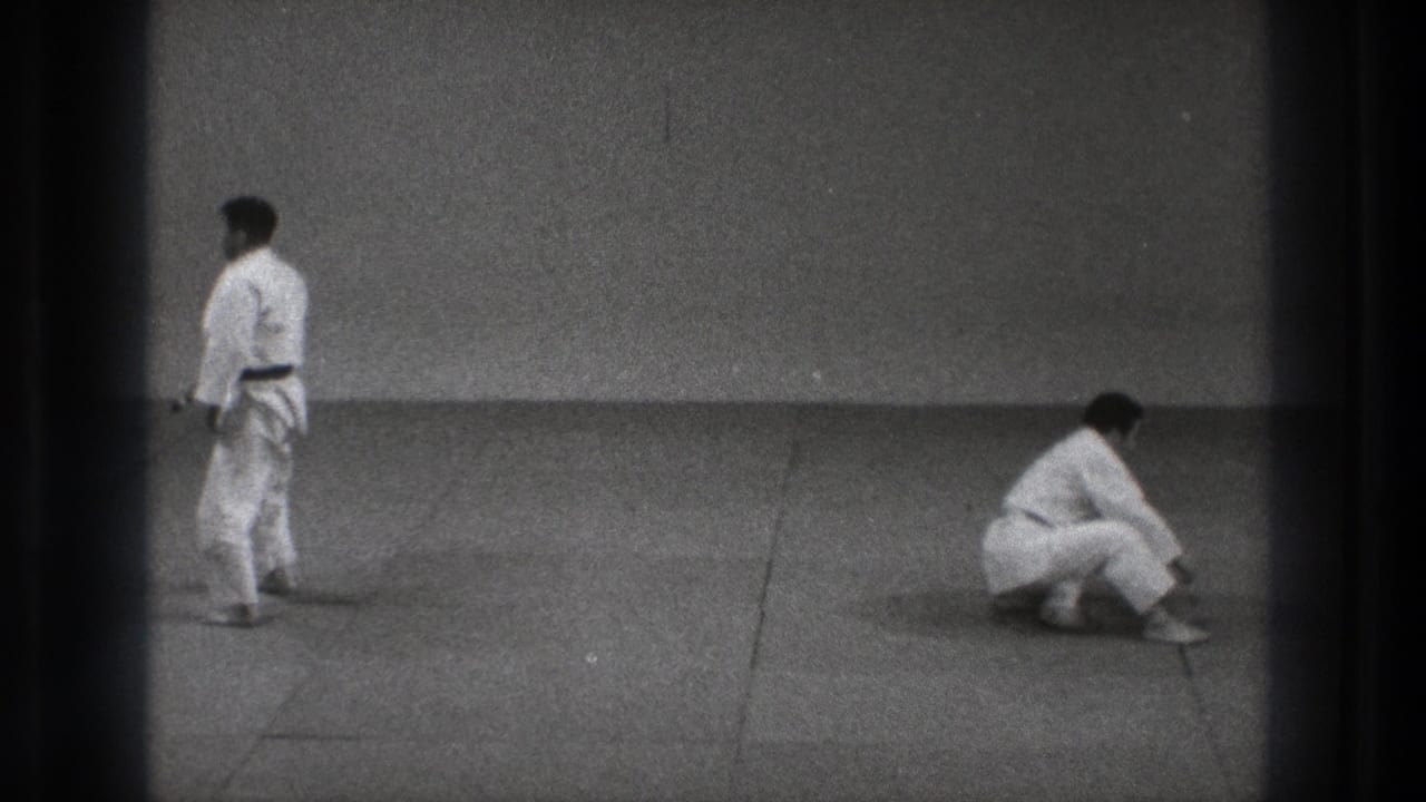 Two karate practitioners in white gi on a mat, one standing and one kneeling low.
