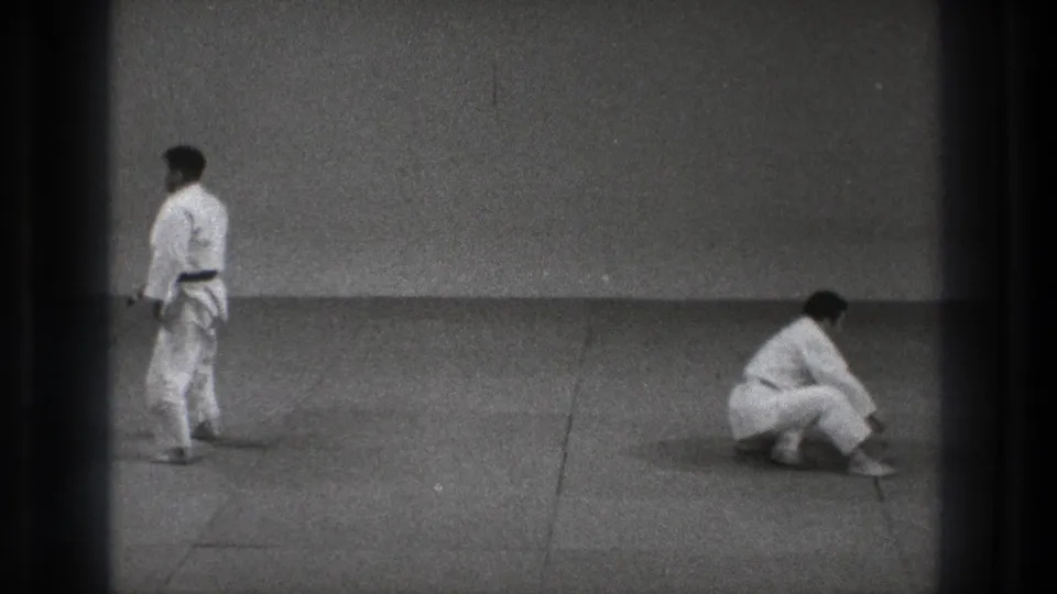 1971 Tokyo 8mm frame showing two adult karate practitioners in white gi on a dojo mat, one upright while the other lowers toward the floor.