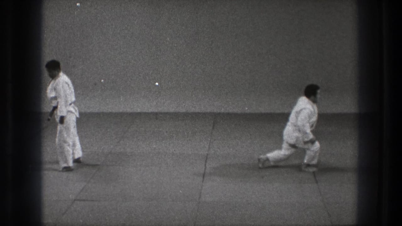 1971 Tokyo dojo frame with one adult practitioner in a low kneel and another standing in a white gi, filmed in grainy black-and-white 8mm.