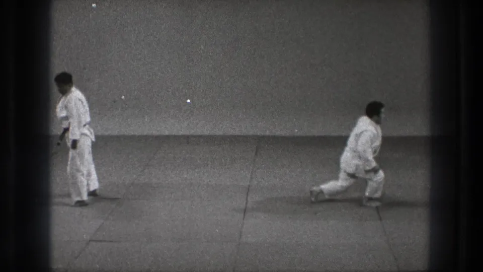 1971 Tokyo dojo frame with one adult practitioner in a low kneel and another standing in a white gi, filmed in grainy black-and-white 8mm.