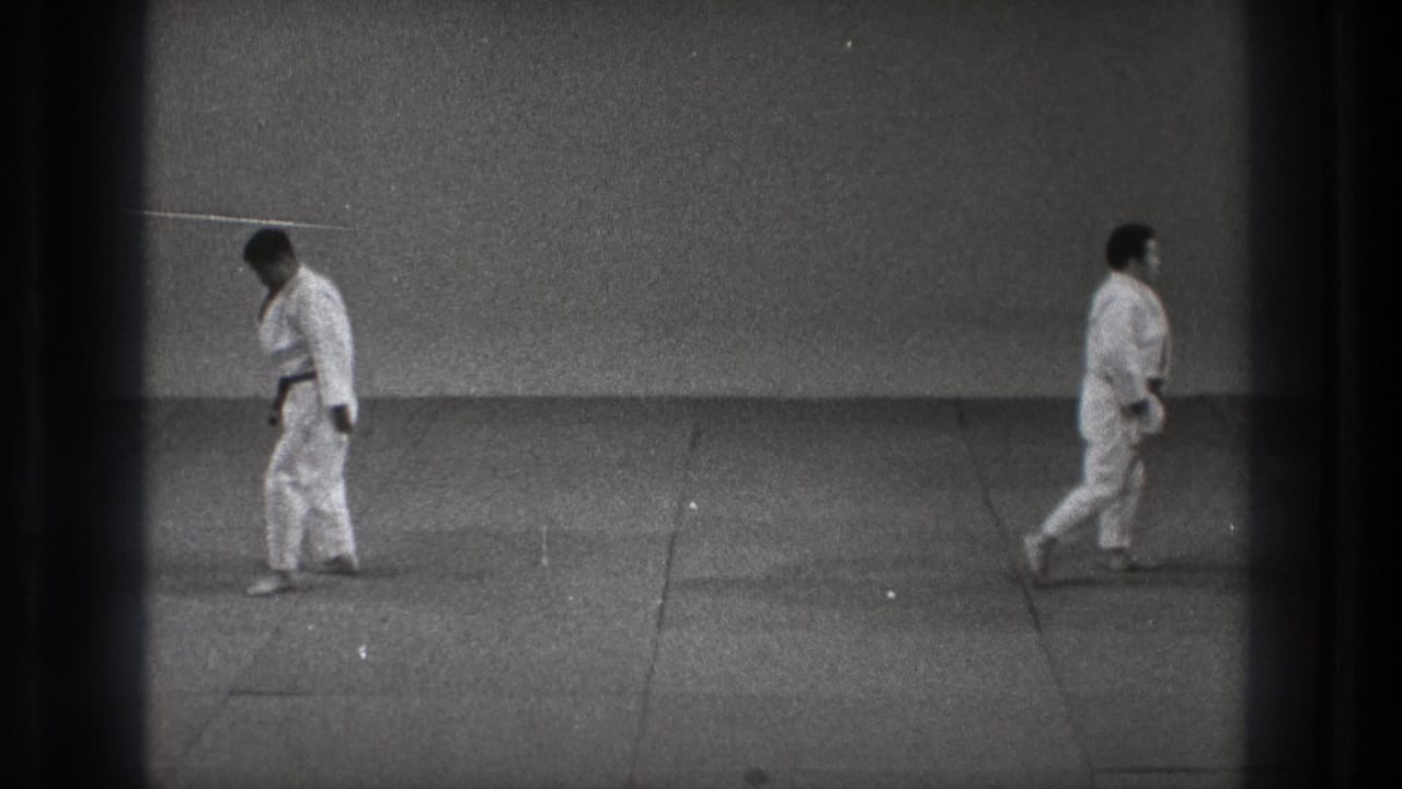 1971 Tokyo training-room frame showing two adult karate practitioners in white gi standing apart on a mat after the movement sequence.