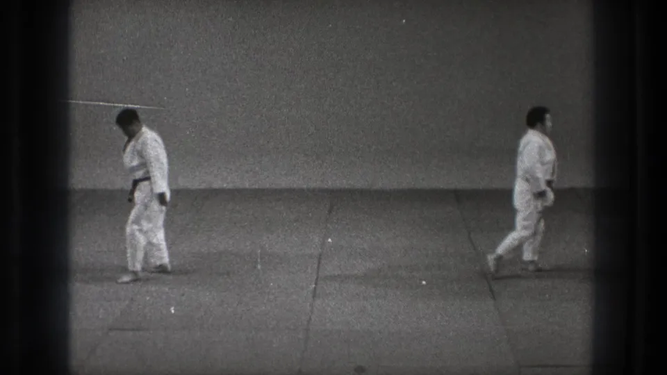 1971 Tokyo training-room frame showing two adult karate practitioners in white gi standing apart on a mat after the movement sequence.