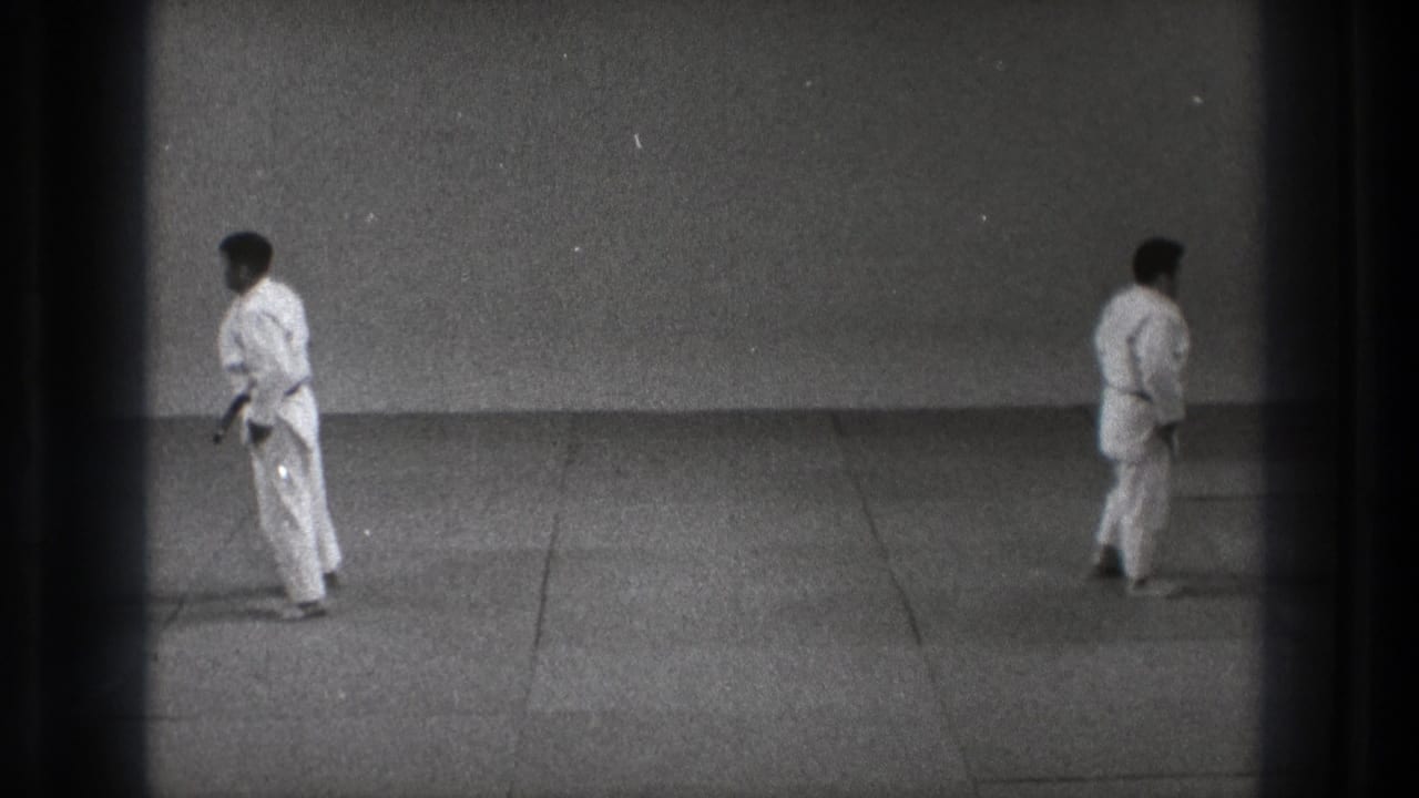 1971 Tokyo 8mm frame showing two adult karate practitioners standing apart in a plain dojo space, with visible film edge darkening.