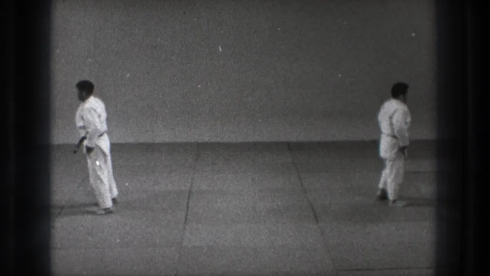 1971 Tokyo 8mm frame showing two adult karate practitioners standing apart in a plain dojo space, with visible film edge darkening.